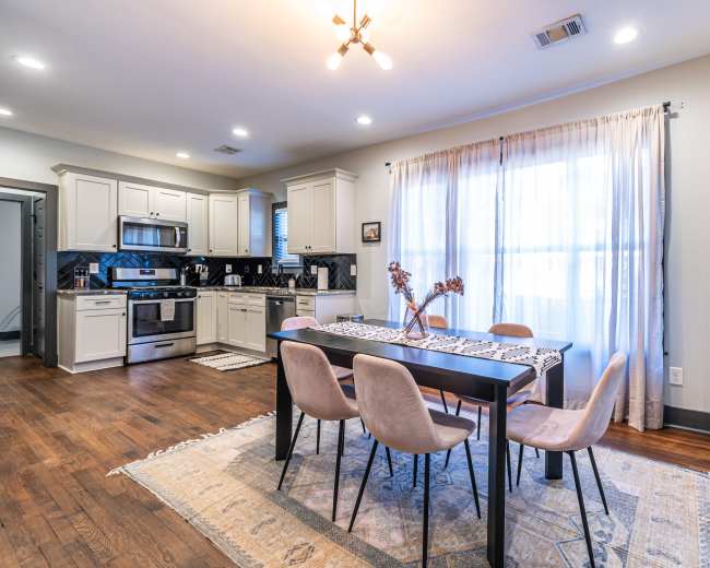 The image shows a modern kitchen and dining area featuring a table with four chairs, a kitchen with gray cabinets, and large windows allowing natural light.