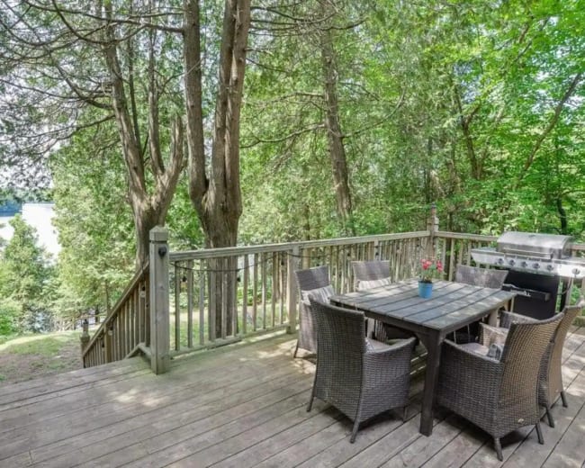A wooden deck is surrounded by trees and features a dining table with chairs and a barbecue grill overlooking a body of water.