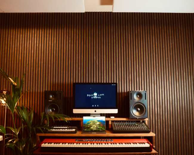 A music production studio features a computer monitor, speakers, a keyboard, and various music equipment against a wooden wall.