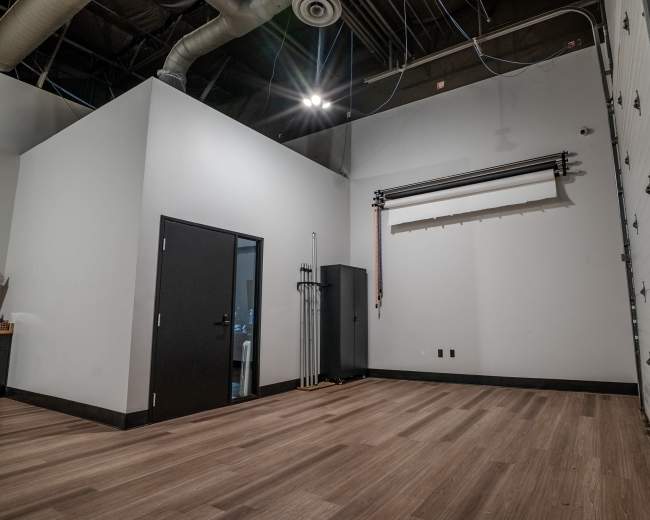 The image shows a spacious interior room with a door on the left, a roll-up door on the right, and wooden flooring.