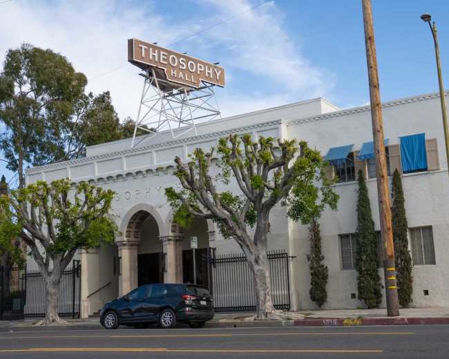 The image shows a white building with arched entrances labeled "Theosophy Hall," flanked by trimmed trees and blue window coverings, under a clear sky.