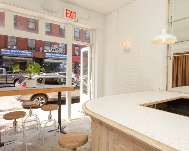 The image shows a bright interior of a cafe with a curved counter, wooden bar stools, and large windows overlooking a street with shops.