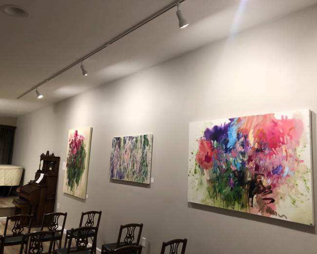 A gallery wall displaying three colorful abstract paintings, with wooden chairs arranged in front.