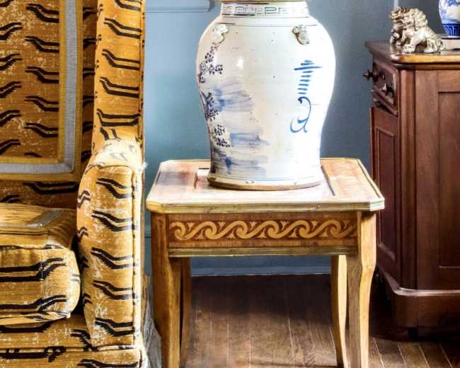 A decorative ceramic jar stands on a wooden side table next to a patterned armchair in a room with wooden flooring and light blue walls.