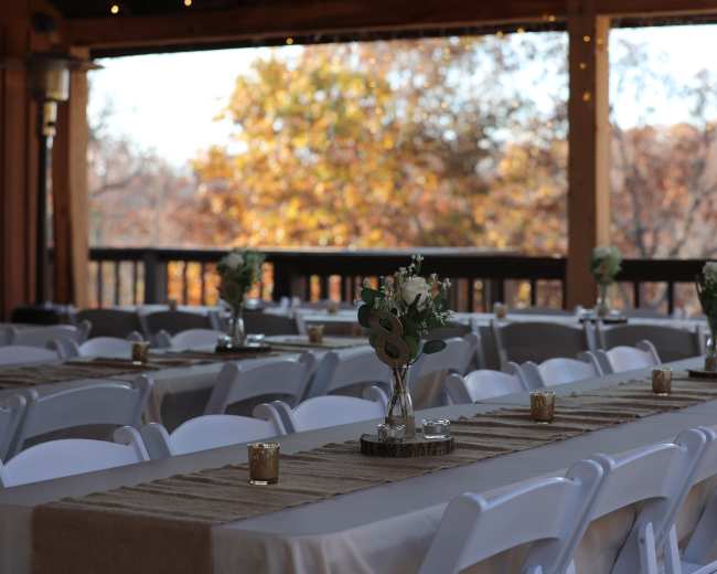 The image shows a banquet-style setup with long tables covered in beige linens, decorated with small vases of flowers and candles, under a wooden pavilion with autumn foliage in the background.