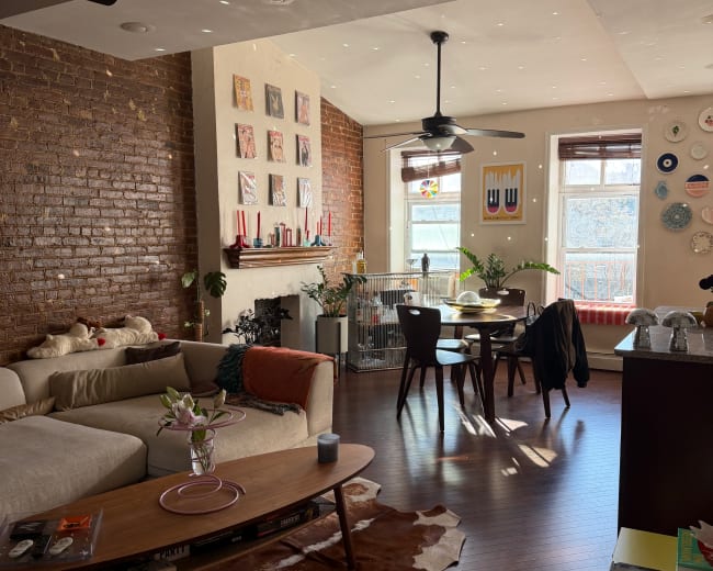 The image shows a bright and modern living space with exposed brick walls, a sectional sofa, a wooden coffee table, and a dining area with large windows.