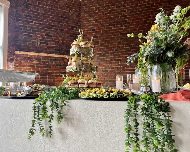 A decorative food display featuring a multi-tiered platter of sandwiches, surrounded by various appetizers and candles, on a table adorned with greenery against a brick wall.