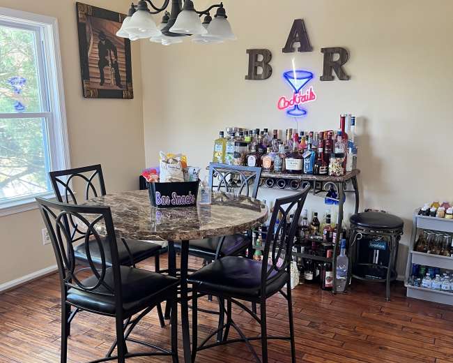 A dining area features a round marble table with four black chairs and a bar cart stocked with various bottles and a neon sign above.