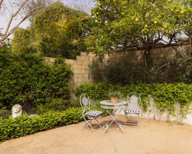 A small patio with a round metal table and two chairs is surrounded by lush greenery and a wall adorned with a lemon tree.