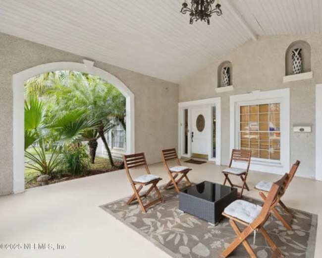 The image shows a covered porch area furnished with six wooden chairs arranged around a small table on a patterned rug.