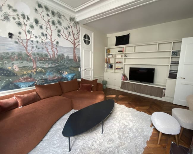 The living room features a curved brown sofa, a black coffee table, white fluffy rug, and a mural of trees and a landscape on one wall.