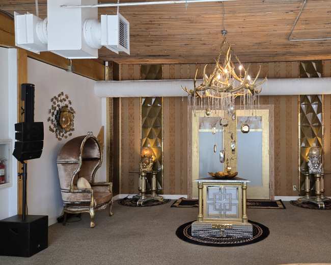 The image depicts a lavishly decorated interior featuring elegant furnishings, including two high-backed chairs, a central ornate table, and a decorative chandelier under wooden ceilings.