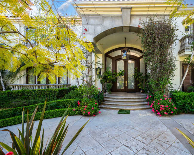 The image shows a grand entrance of a house with a curved stone staircase, large wooden double doors, and lush greenery surrounding the pathway.