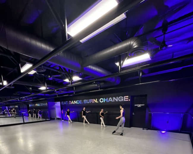 A group of dancers practices in a spacious studio with black walls and purple lighting.
