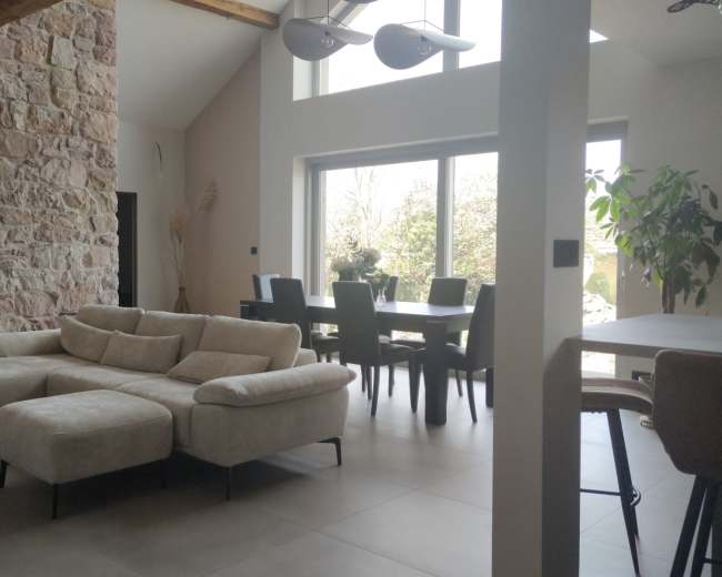 The image shows a modern living room with a light-colored sectional sofa, stone walls, and a dining area featuring a large table and dark chairs under bright natural light from large windows.