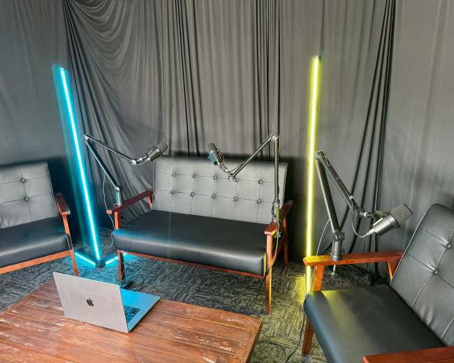 A recording setup with two black leather couches, two adjustable microphones on stands, and a laptop on a wooden table, all set against a backdrop of dark fabric.
