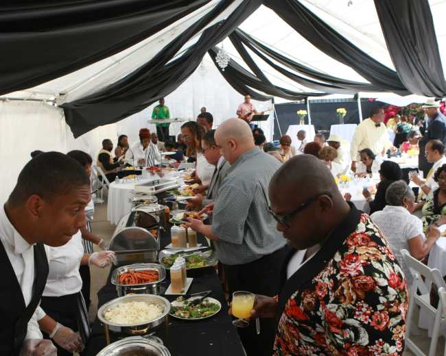 A catered event is taking place under a tent, with guests serving themselves food from a buffet while others are seated at white tables.