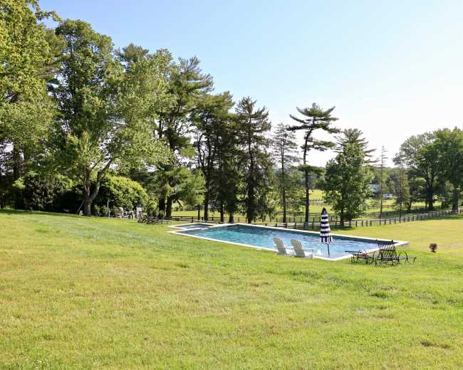 A swimming pool is situated on a grassy area surrounded by trees and a fenced pasture.