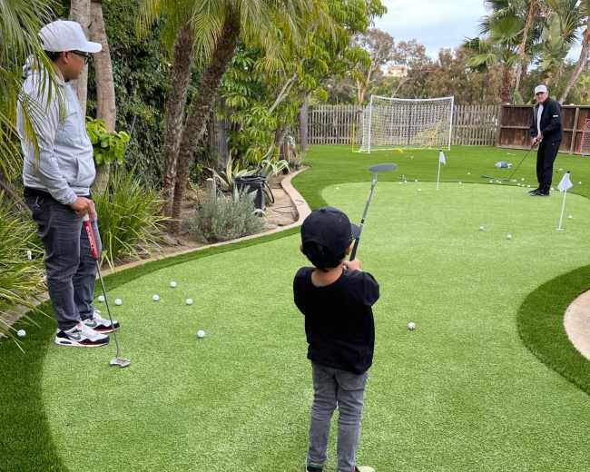 A child practices putting on a mini-golf green in a backyard while an adult observes.
