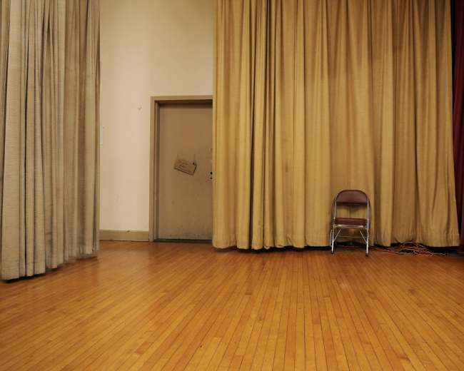 An empty stage features a single chair against a backdrop of golden curtains and a door.
