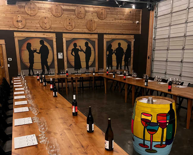 A large wooden dinner table is arranged in a U-shape with wine glasses and bottles, surrounded by silhouetted figures on the walls and a colorful painted barrel at one end.