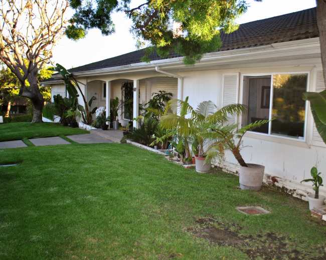 A single-story house with a green lawn, bordered by a walkway and surrounded by various potted plants along the side.