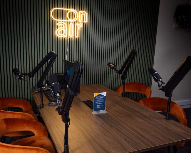 A modern podcast studio features a wooden table with four microphones and several orange chairs, illuminated by a neon "on air" sign on the wall.