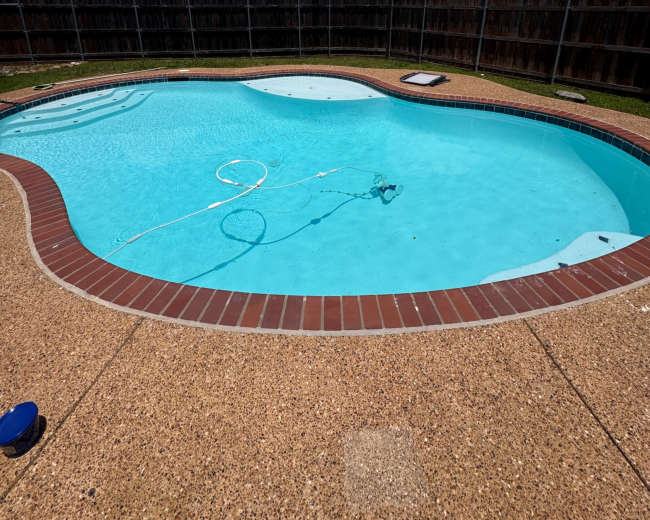 A clear blue swimming pool is surrounded by a brown brick patio and a wooden fence under a partly cloudy sky.