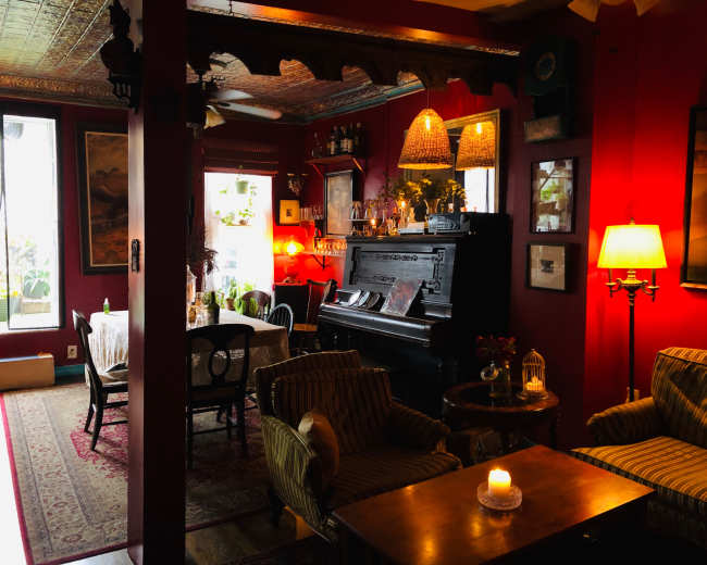 A cozy lounge area features a piano, vintage furniture, and warm lighting, with plants visible through large windows.