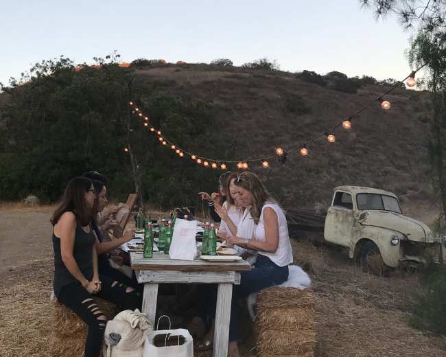 A group of people sits around a rustic table with drinks and food, surrounded by hay bales and string lights in an outdoor setting.