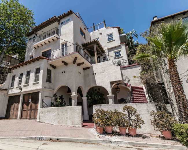 The image shows a three-story stucco house with arched doorways, a red-tiled roof, and a driveway lined with potted plants.