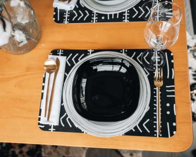 A dining table set with a black square bowl, a round white plate, a gold spoon, a glass of water, and patterned placemats.