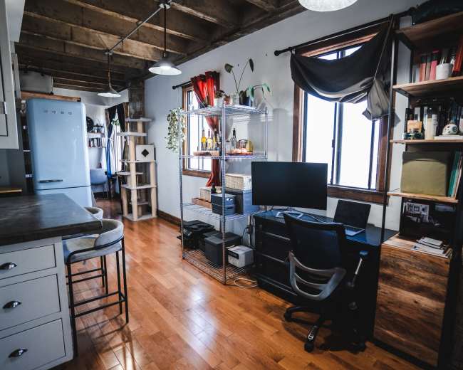 A home office setup features a desk with a computer monitor and shelves filled with books and plants, set against wooden flooring and large windows.