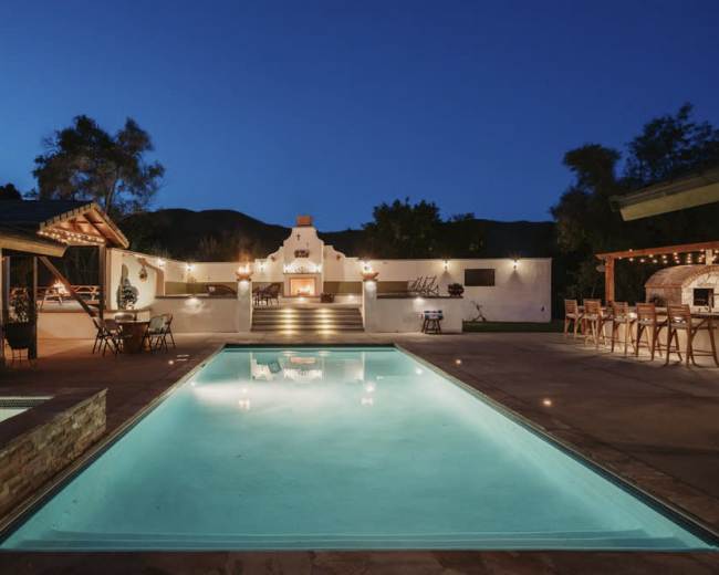 A well-lit outdoor area featuring a swimming pool, a barbecue space, and various seating arrangements in a nighttime setting.