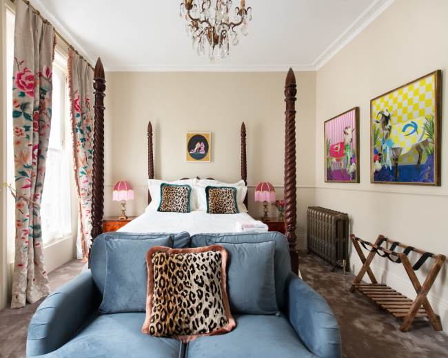 A bedroom features a king-size bed with tall wooden posts, a couch with leopard-print pillows, and colorful artwork on the walls.