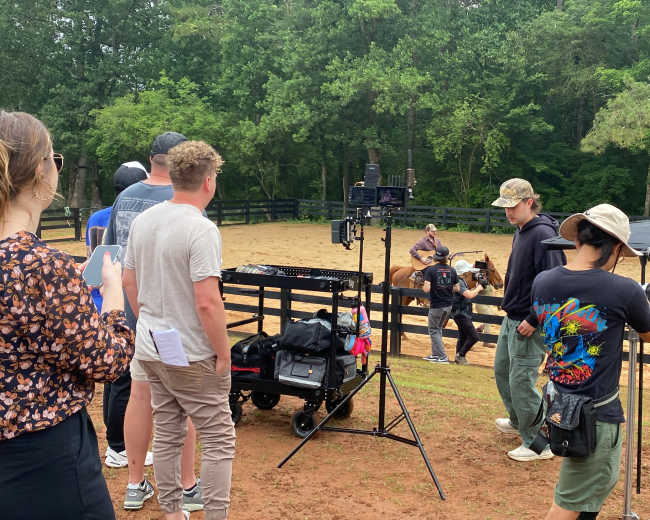 A group of people is gathered near a filming setup in an outdoor area with a horse and a rider in the background.