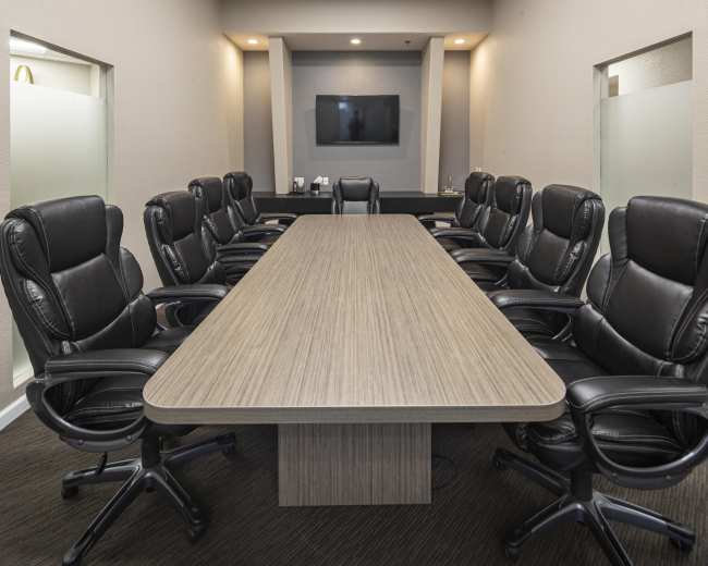 A conference room features a long wooden table surrounded by several black leather office chairs, with a television mounted on the wall at one end.