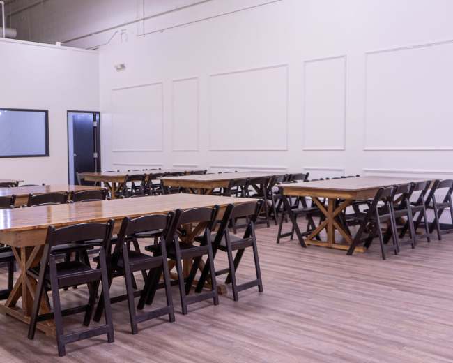 The image shows a spacious room with several rectangular wooden tables and black folding chairs arranged in a grid pattern, set against plain white walls.