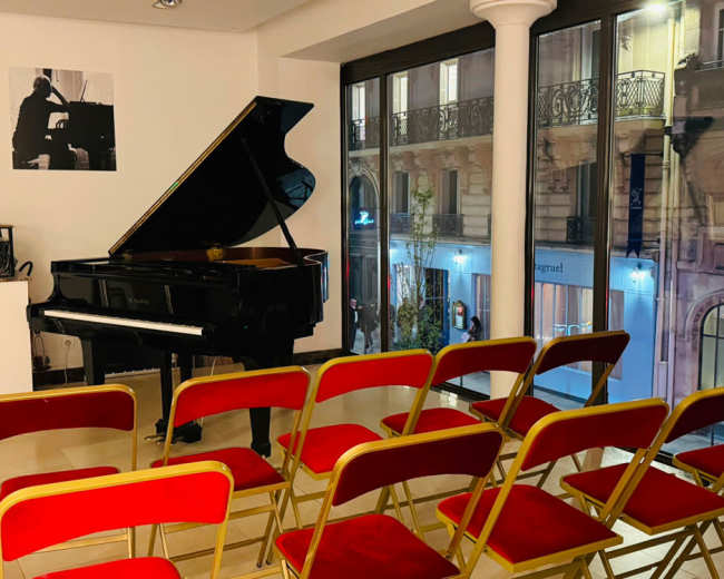 The image shows a grand piano positioned near a large window, with several red chairs arranged in front of it in a sparsely decorated room.