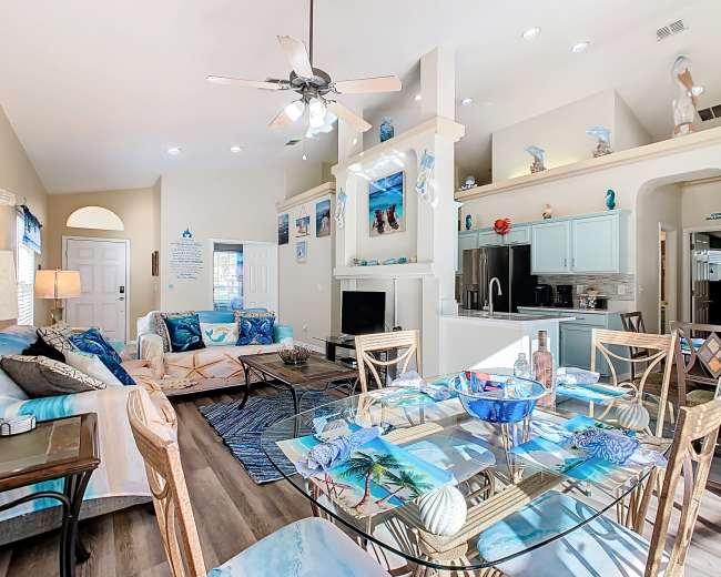 The image shows a bright, open living area with a dining table, comfortable seating, and light blue kitchen cabinets.