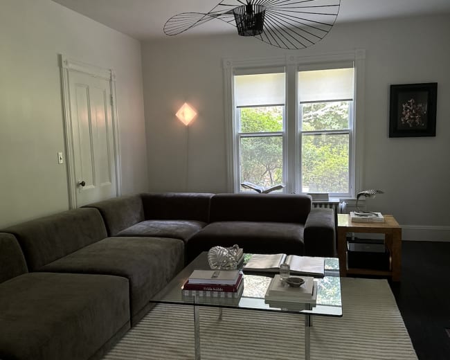 The image shows a living room with a large sectional sofa, a glass coffee table, and natural light coming through the windows with a view of greenery outside.