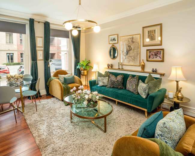 The image shows a well-decorated living room with a green velvet sofa, a round glass coffee table, and a combination of armchairs and dining chairs arranged near a window.