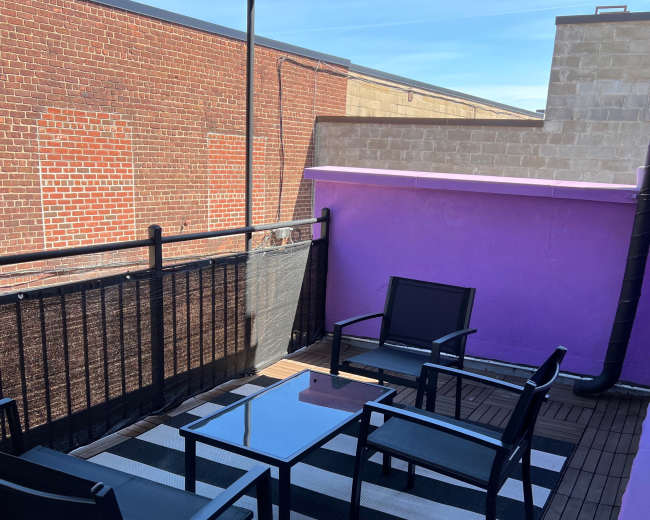 The image shows a rooftop terrace with a striped rug, a glass table, and black chairs against a backdrop of brick and purple walls.