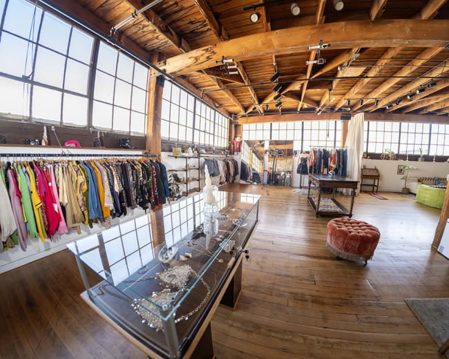 The image shows a spacious clothing store with wooden floors, featuring racks of colorful apparel and a display case with jewelry in the center.