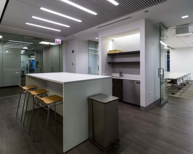 The image shows a modern office kitchen area featuring a long white countertop with barstools, a dishwasher, and glass-paneled meeting rooms in the background.