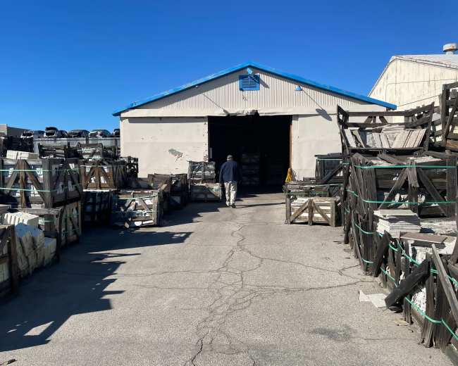 A person walks towards a large storage building surrounded by stacked crates and containers under a clear blue sky.