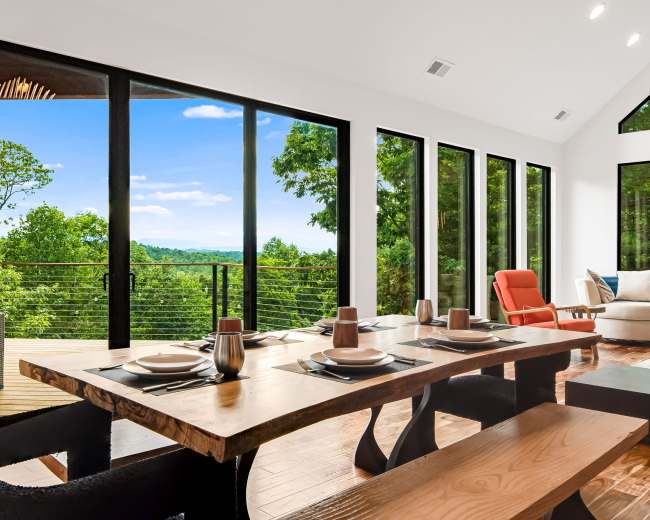 A modern dining area features a wooden table set for a meal, with large glass windows showing a view of lush greenery and a bright sky.