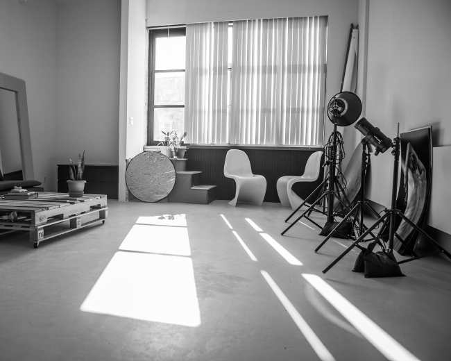 The image shows a minimalist interior space with a wooden pallet sofa, two modern chairs, a circular table, and photography lighting equipment.