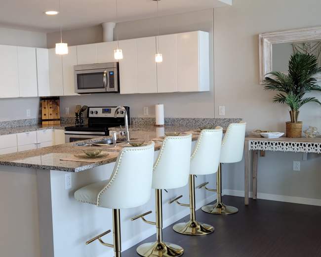 The image depicts a modern kitchen equipped with white cabinets, stainless steel appliances, a granite countertop, and four bar stools.