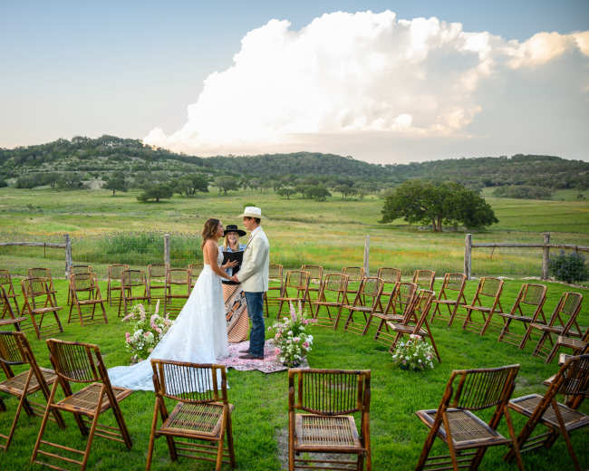 A couple stands in a grassy clearing surrounded by a circle of empty wooden chairs, with rolling hills and clouds in the background.